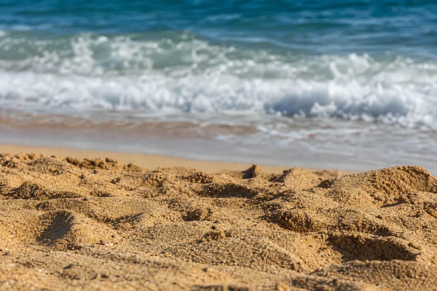 Close-up of coarse golden sand in sharp focus in the foreground, with a foamy breaking wave and blue sea blurred behind