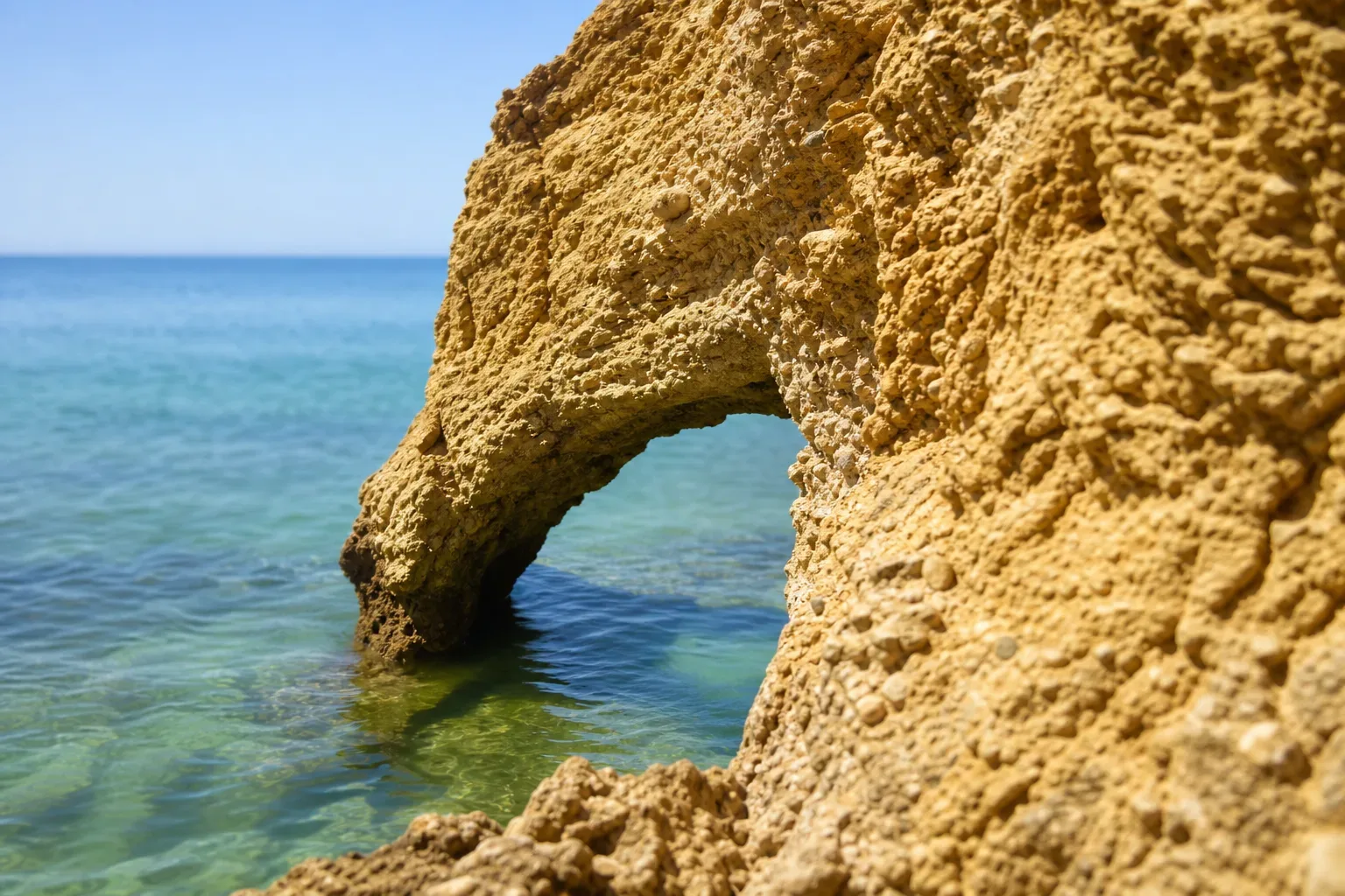 A natural arch in textured golden limestone sits above clear turquoise coastal water beneath a bright blue sky