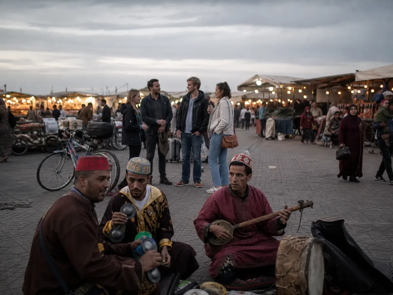 Three street musicians in traditional dress sit on paved ground playing instruments, while a small group of people chat in the middle distance amid market stalls lit by strings of lights at dusk