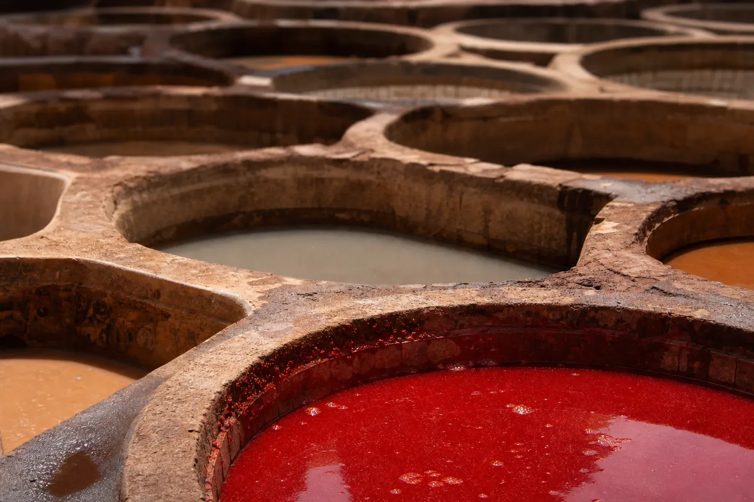 Close-up of circular stone dyeing vats filled with liquids in vivid red and pale grey tones, with further vats receding out of focus behind