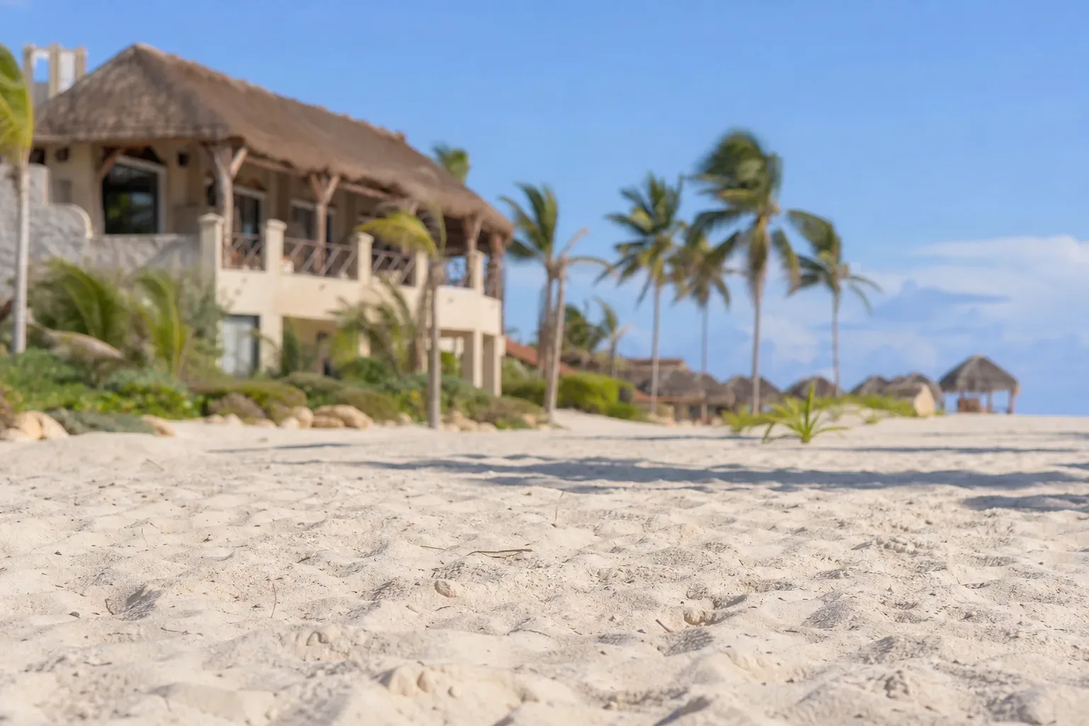 A thatched beachfront resort building with palm trees stands behind empty pale sand shot in sharp close focus with shallow depth of field