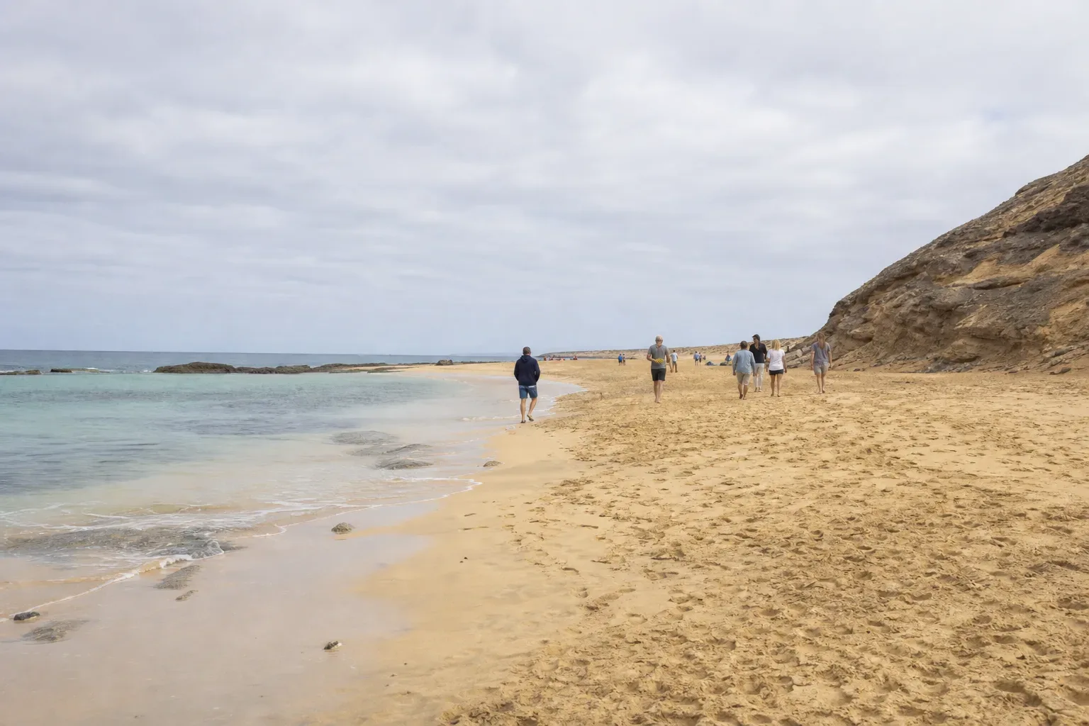 Several people walk along a sandy beach beside calm, pale-turquoise water, with a rocky cliff rising on one side under an overcast sky