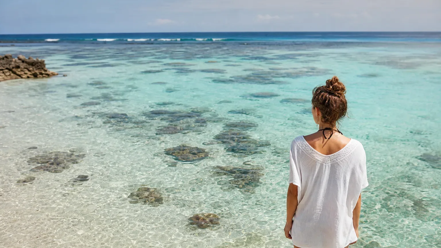 A person in a white top stands at the edge of crystal-clear shallow water over a coral reef, looking out towards the open ocean