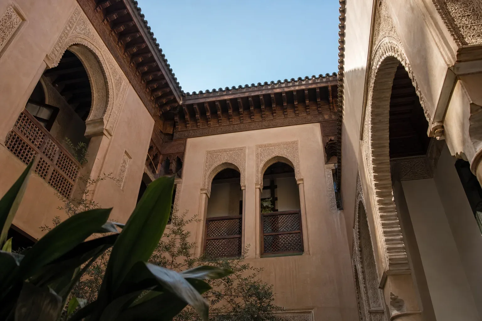 A low-angle view looking up at ornately carved stone archways and latticed wooden screens surrounding an open courtyard, with blue sky visible above