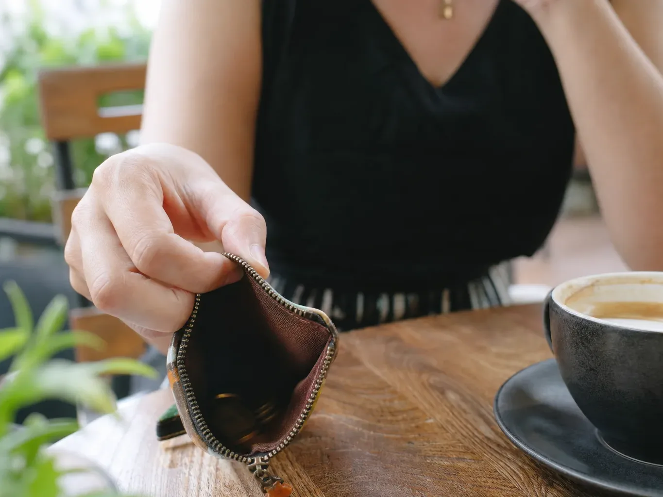 A person sits at a wooden café table, holding open an empty zip coin purse beside a cup of coffee