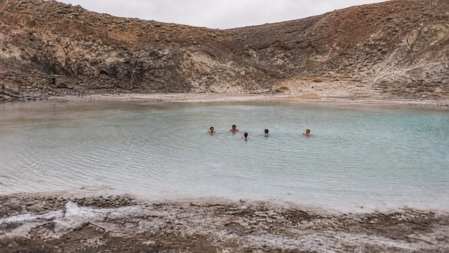 Five people swim in a pale turquoise crater lake surrounded by barren rocky volcanic walls under an overcast sky