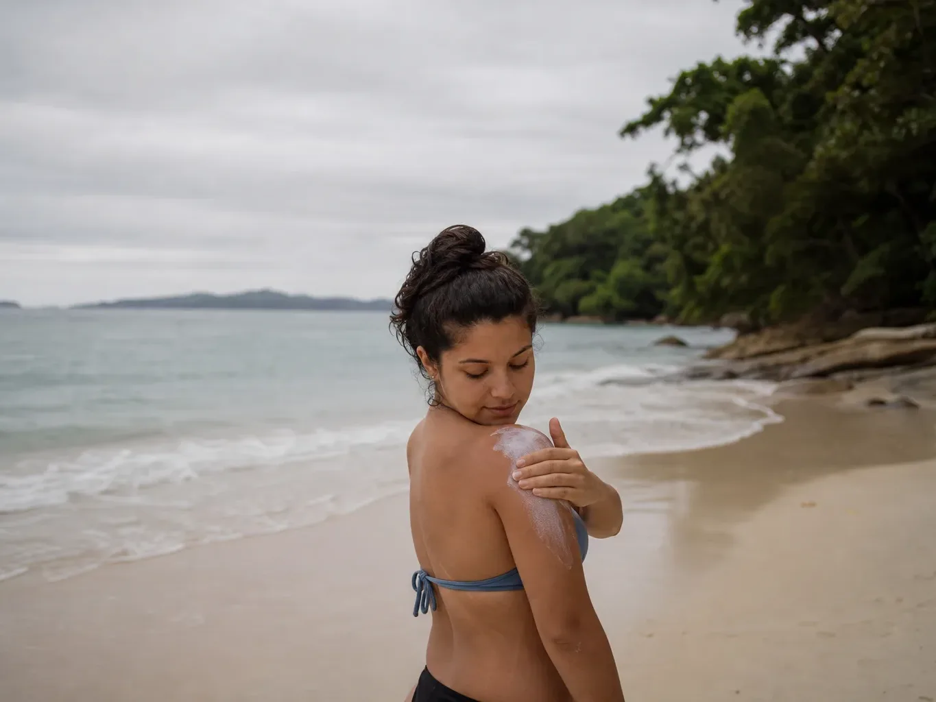 A person in a bikini top applies cream to their shoulder while standing on a sandy beach with calm turquoise water and lush trees behind